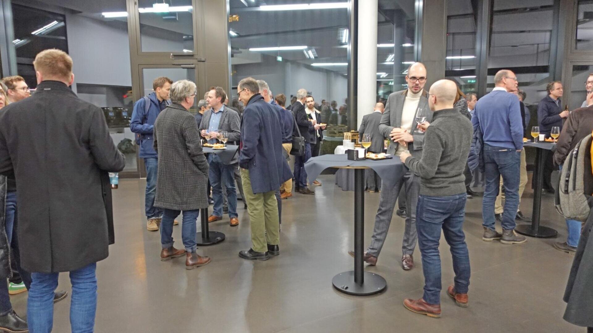 A group of people in business attire stand and talk in small groups at a networking event in a modern indoor space with large windows and high tables. Some hold drinks and snacks.