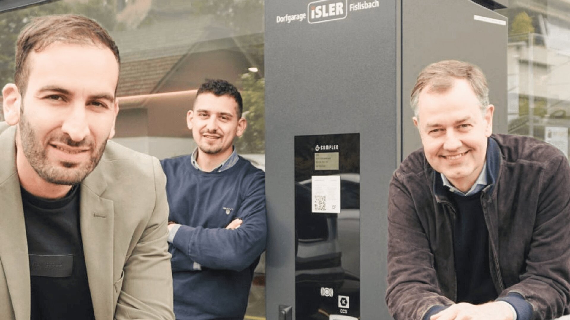 Drei Männer lächeln und posieren neben einer großen grauen elektronischen Ladestation vor einem Gebäude mit dem Schild "Dorfgarage Isler" darüber. Der Hintergrund zeigt ein Fenster, in dem sich Bäume und Dächer spiegeln.