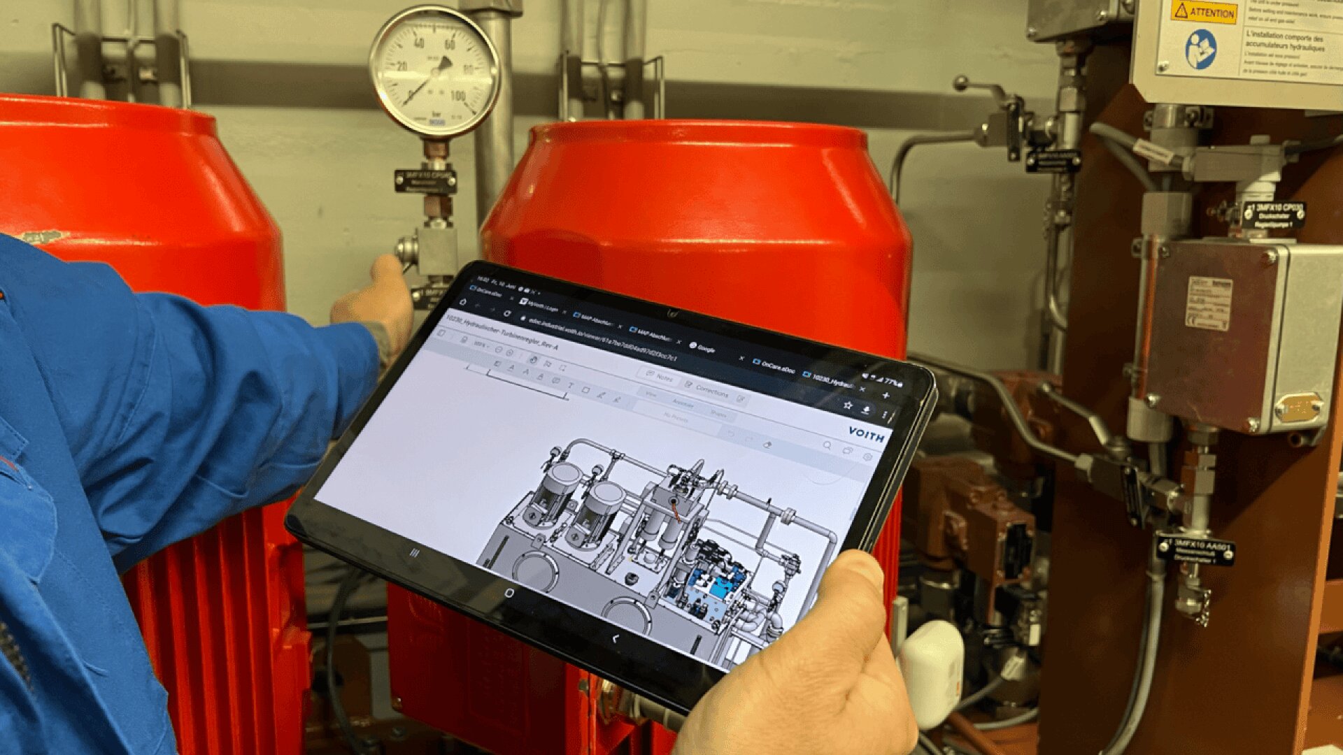 A person holds a tablet displaying a technical diagram in front of industrial machinery with red components, while adjusting a valve near a pressure gauge.