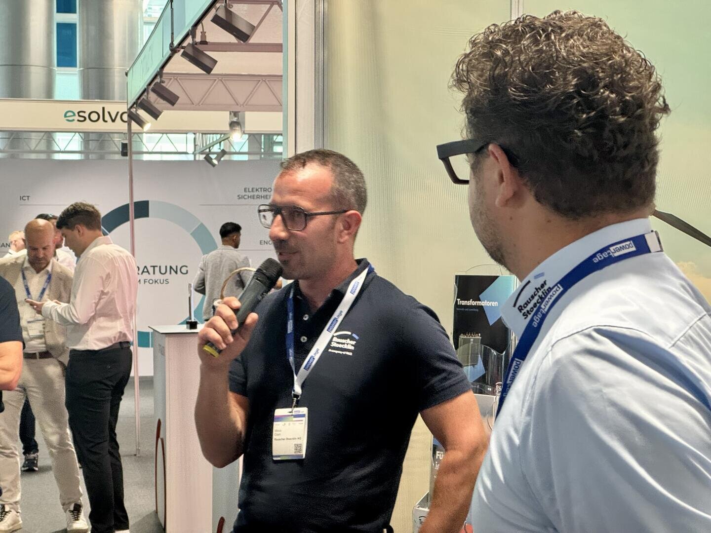Zwei Männer, die Umhängebänder tragen, sprechen auf einer Konferenz oder Messe in Mikrofone. Der eine blickt in die Kamera, der andere ist von der Seite zu sehen. Im Hintergrund sind Stände mit Bannern und Besucher zu sehen.