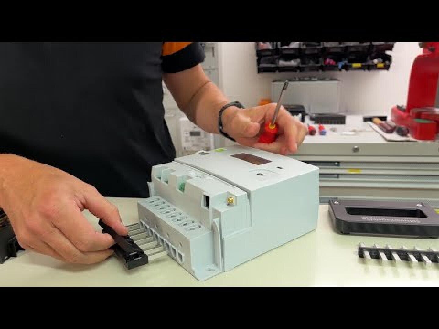 A person uses a screwdriver to assemble or repair an electrical component or circuit breaker on a workbench, with various tools and parts in the background.