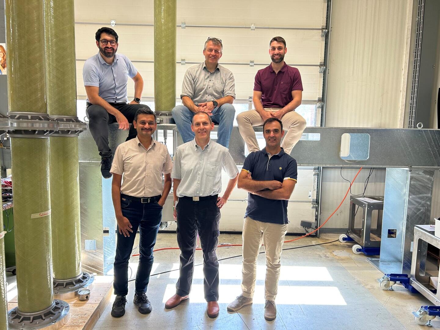 Sechs Männer posieren in einem hellen Industrielabor mit großen strukturellen Säulen und Geräten. Drei stehen vorne, drei sitzen oder hocken auf Balken hinter ihnen. Sie sind leger gekleidet und lächeln in die Kamera.