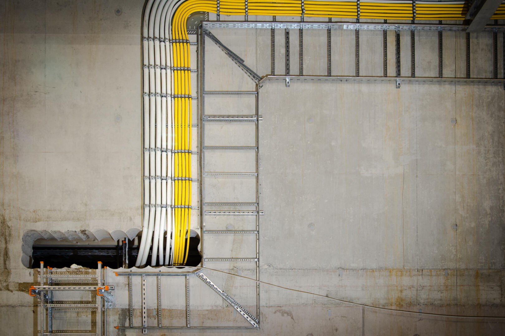 Eine Reihe von gelben und weißen Kabeln, die ordentlich auf metallenen Kabelrinnen angeordnet sind, verläuft entlang einer Betonwand und -decke und macht dabei scharfe rechtwinklige Kurven. Die Kabelrinnen werden von Metallbügeln gehalten.