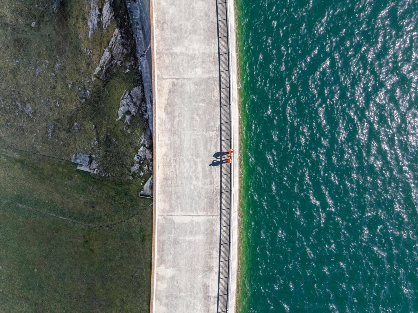 Luftaufnahme einer Person, die auf einer breiten Betonbrücke mit Metallgeländer steht, die eine Grasfläche mit Felsen auf der linken Seite und tiefgrünem Wasser auf der rechten Seite trennt.