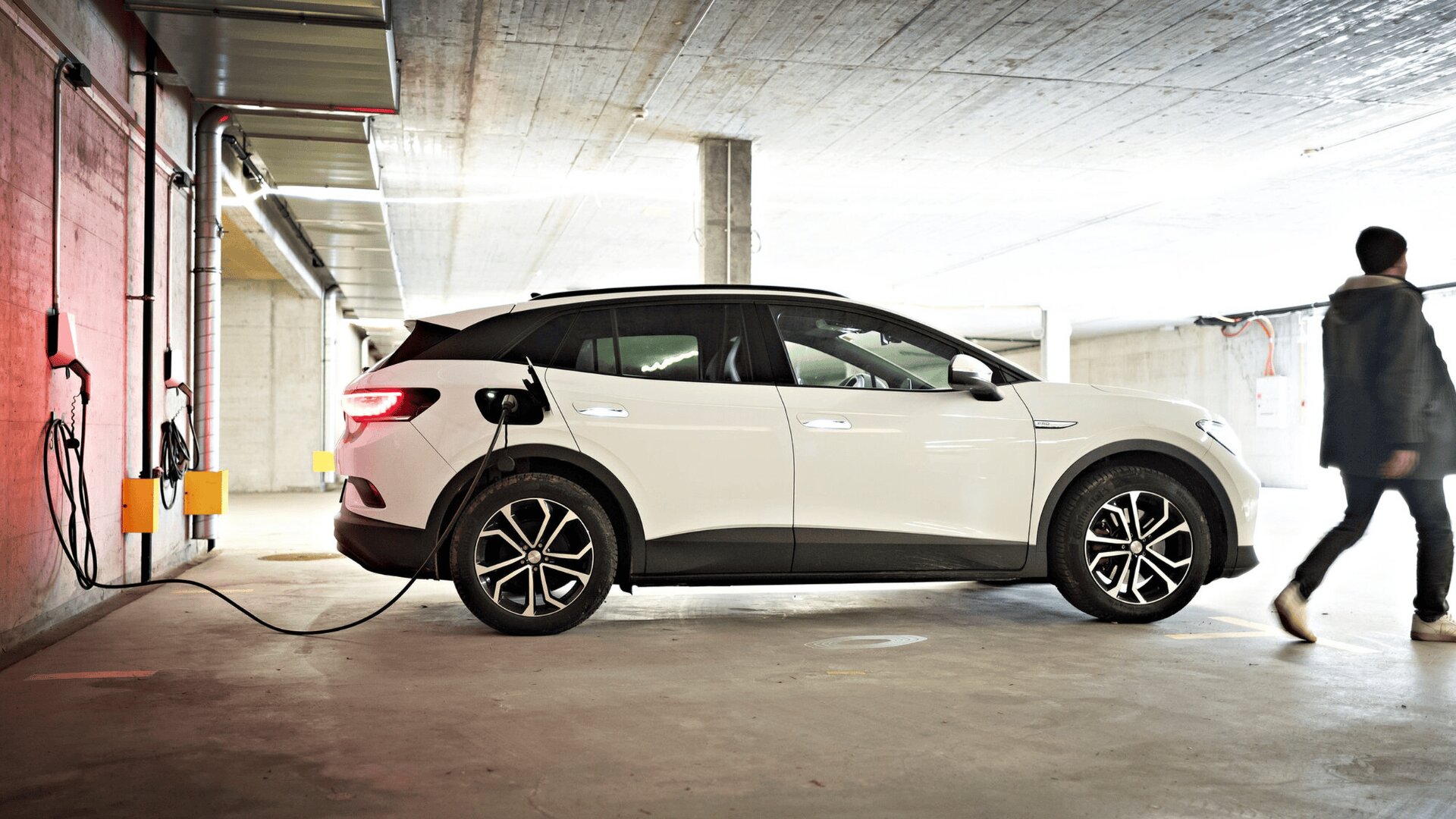 Ein weißer Elektro-Geländewagen ist in einer Garage geparkt und an eine Ladestation angeschlossen. Auf der rechten Seite des Bildes geht eine Person vorbei.