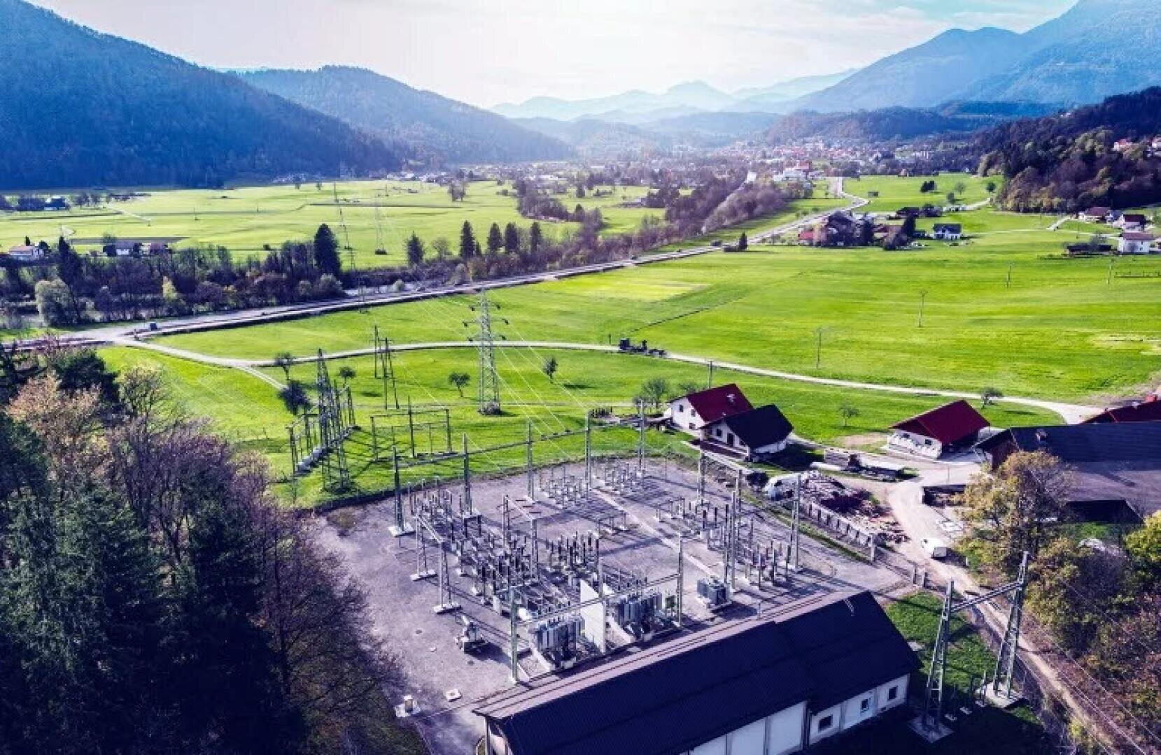 Luftaufnahme eines Umspannwerks, umgeben von grünen Feldern, Häusern und Bäumen, mit Bergen und einer kleinen Stadt in der Ferne unter einem klaren, hellen Himmel.