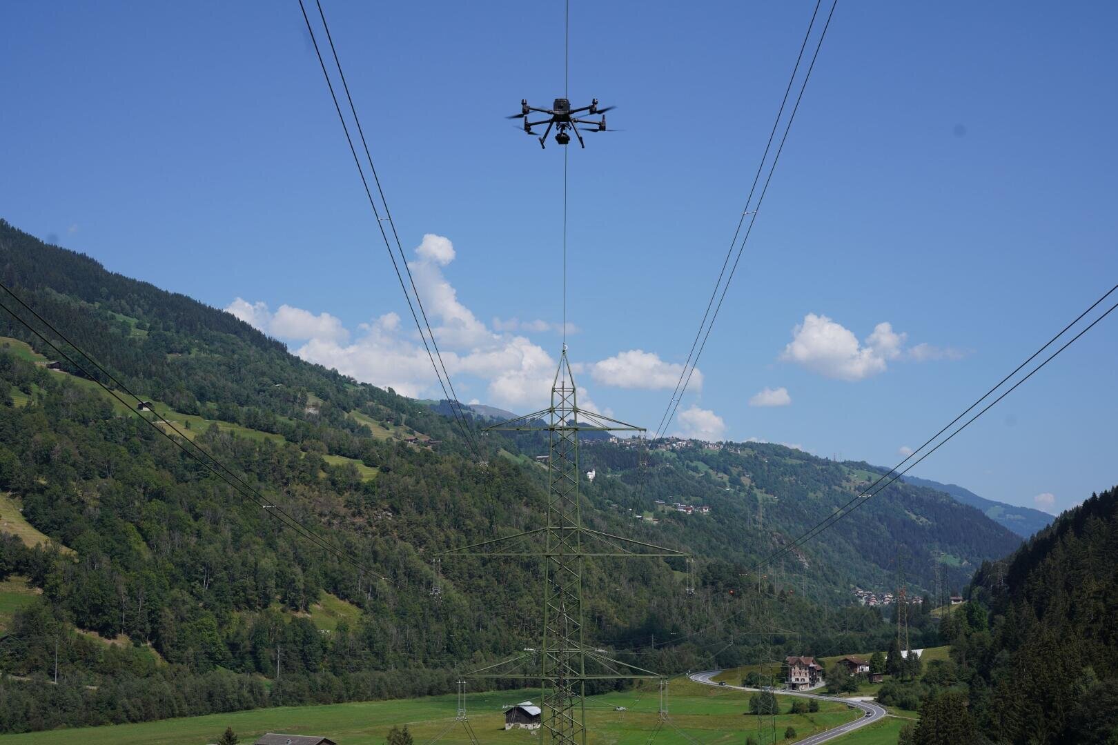 Eine Drohne schwebt über Stromleitungen und einem Sendemast in einem grünen Gebirgstal, mit einer kurvenreichen Straße und verstreuten Häusern unter einem klaren blauen Himmel.