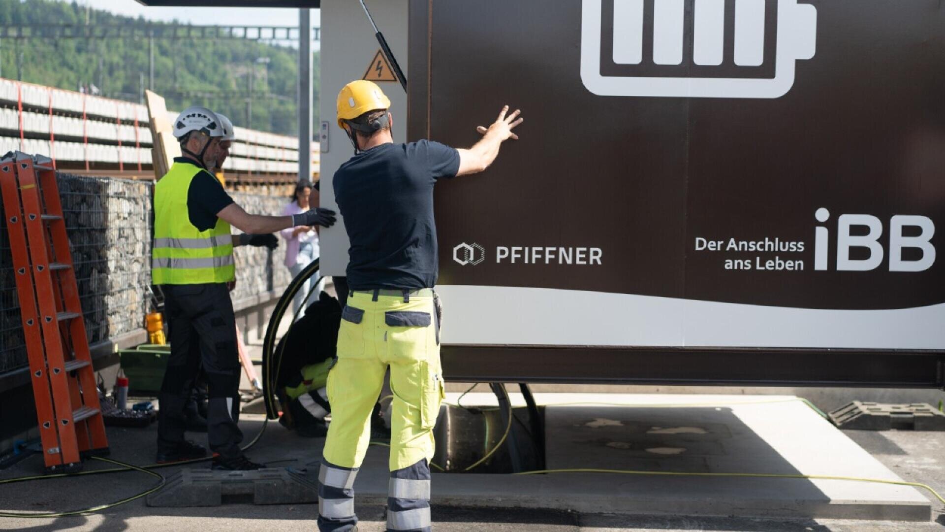 Two construction workers in safety gear stand near a large brown container with a battery icon and logos. One worker gestures toward the container, while the other handles equipment next to a ladder and stone wall.