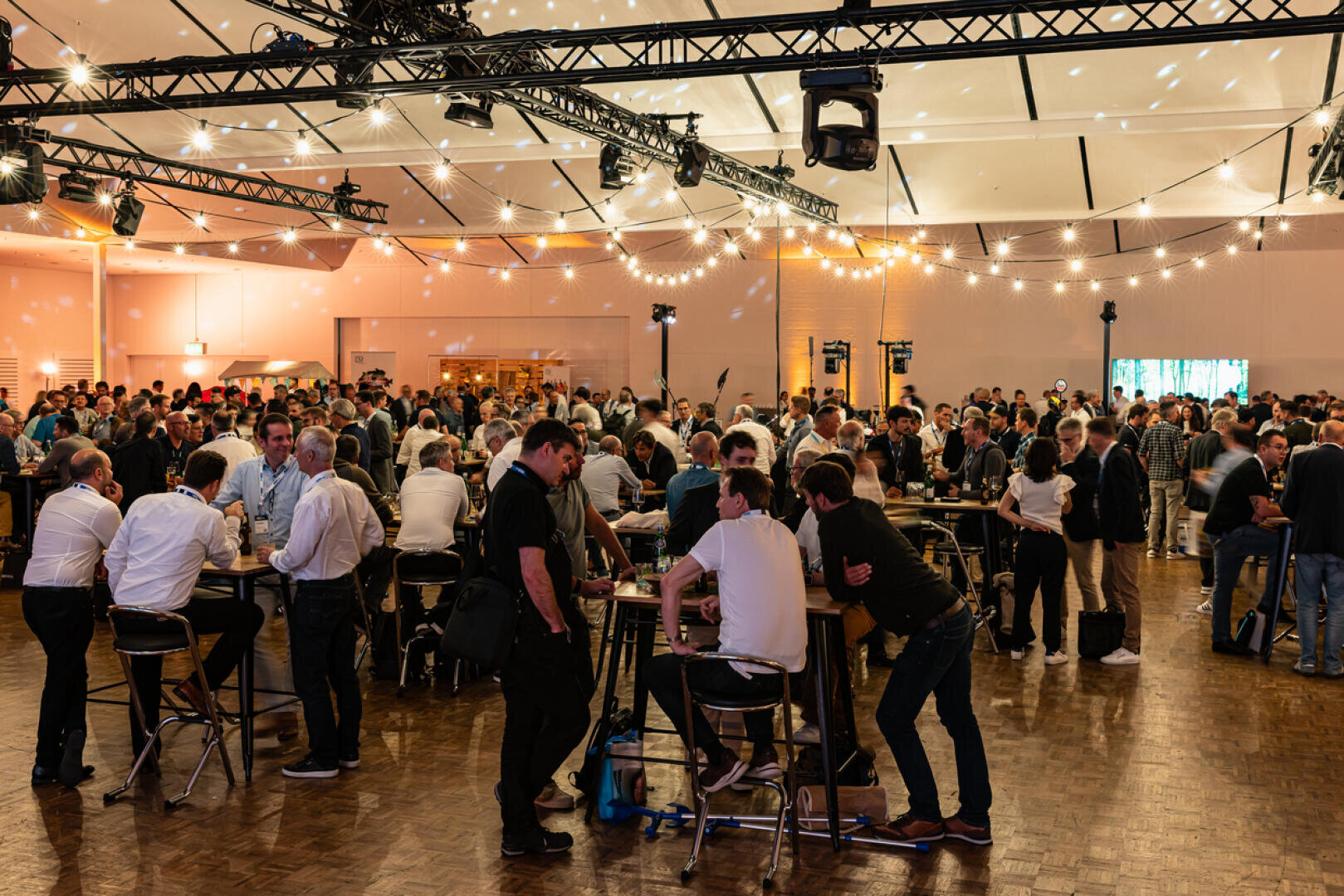 A large group of people gather in a spacious, well-lit hall, chatting and meeting at high tables. Fairy lights and ceiling lights create a warm, festive atmosphere.