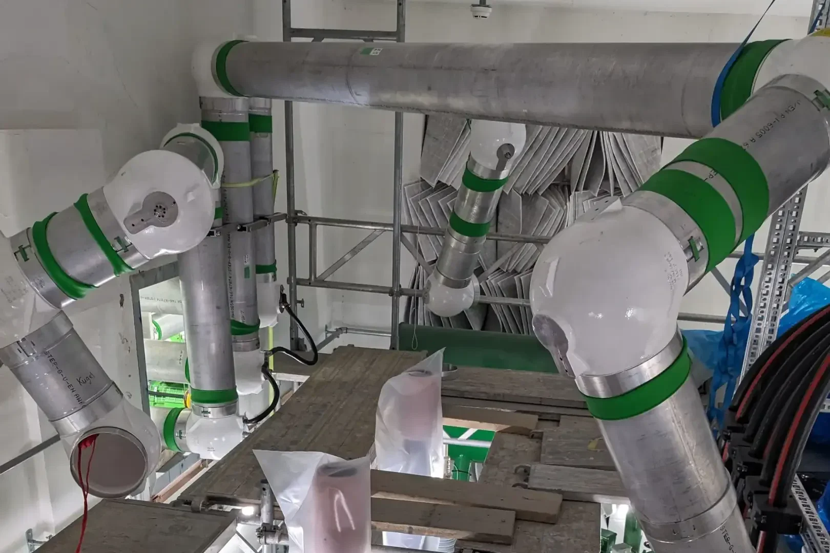 Industrierohre mit weißen und grünen Markierungen verlaufen durch eine Baustelle, die von Gerüsten und Plattformen umgeben ist. Einige Rohre haben abgewinkelte Muffen, und Säcke oder Behälter werden auf Holzbrettern darunter abgestellt.