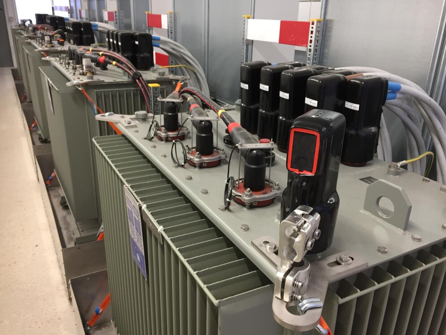 Eine Reihe großer industrieller elektrischer Transformatoren mit grauen gerippten Gehäusen und schwarzen oberen Anschlüssen, die durch dicke Kabel verbunden sind.