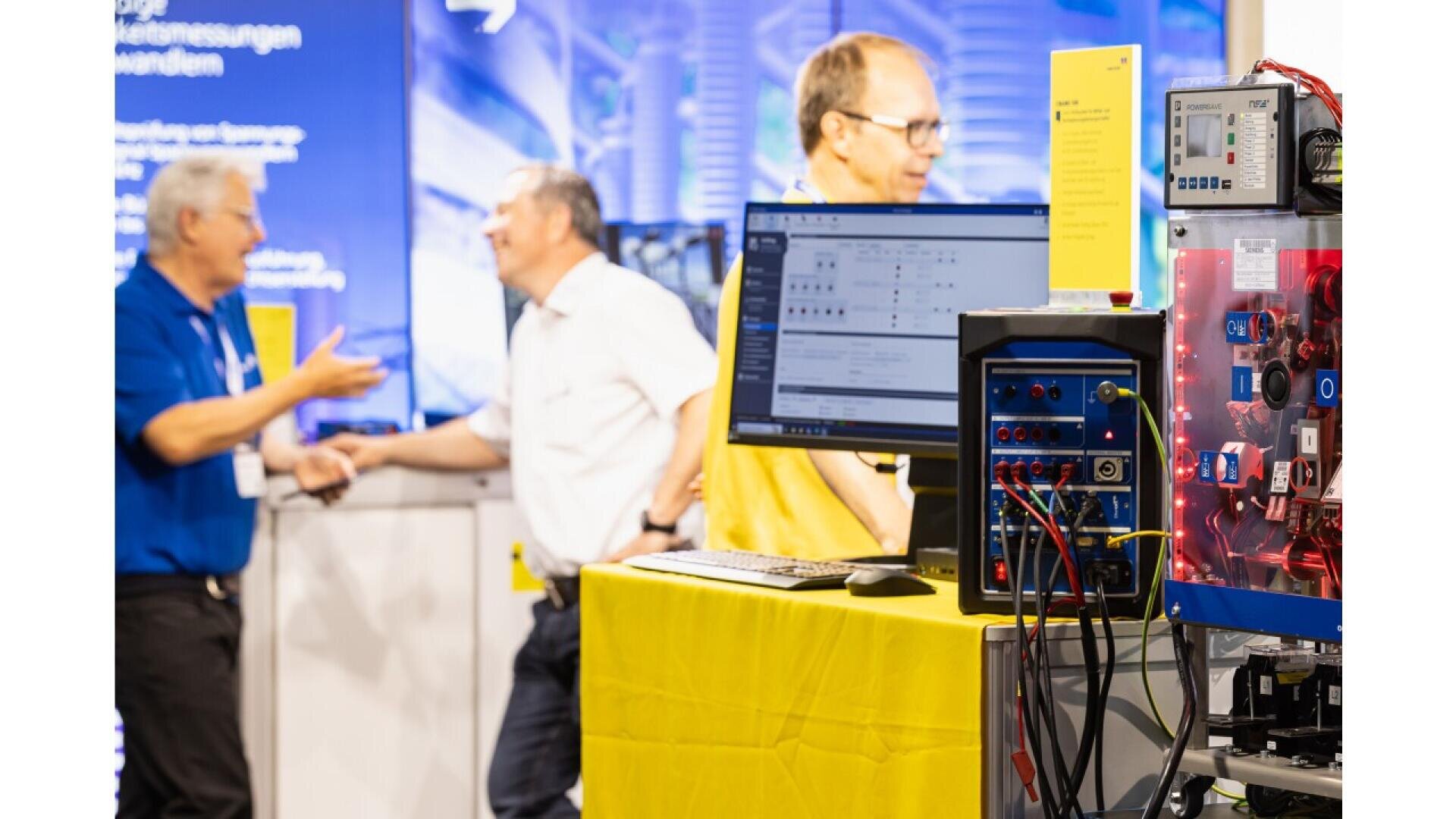 A computer workstation with electronic equipment is shown in the foreground, while three men converse and smile in the blurred background at a technology exhibition or trade show.