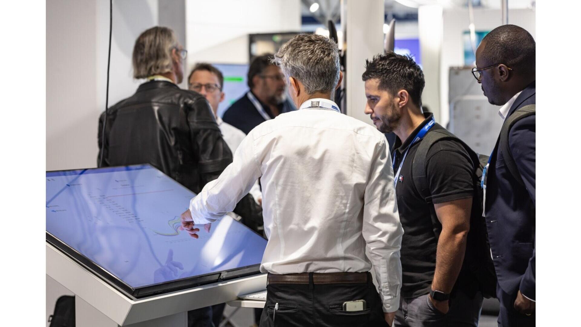 A group of men gathers around a large touchscreen display; one man points at the screen while others watch attentively, suggesting a collaborative discussion or demonstration.