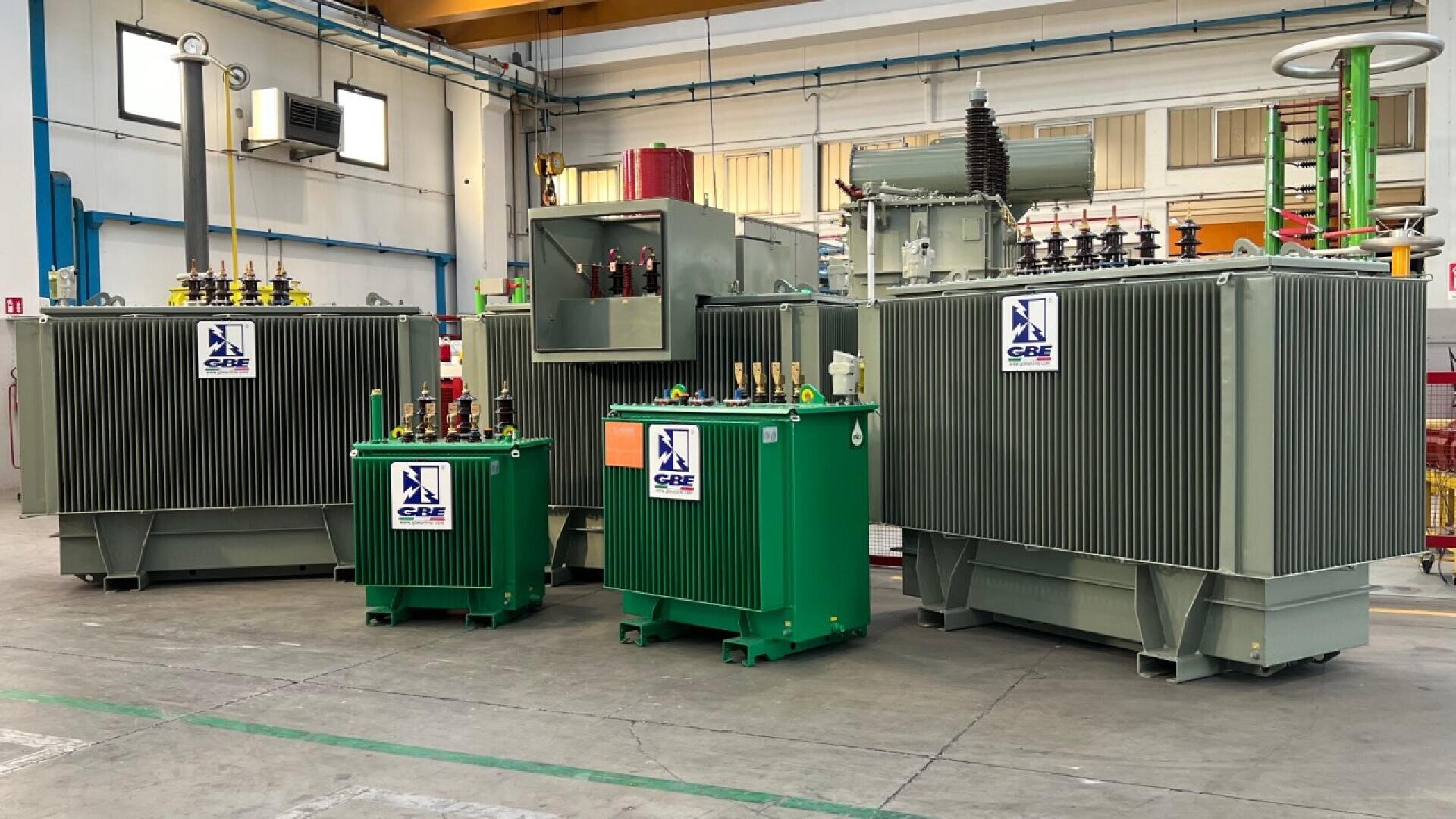 Drei große elektrische Transformatoren, zwei graue und ein grüner, stehen in einer Industrieanlage. Alle tragen das CBE-Logo, und oben sind verschiedene Kabel und isolierte Klemmen zu sehen. Im Hintergrund sind Fenster und Industrieanlagen zu sehen.