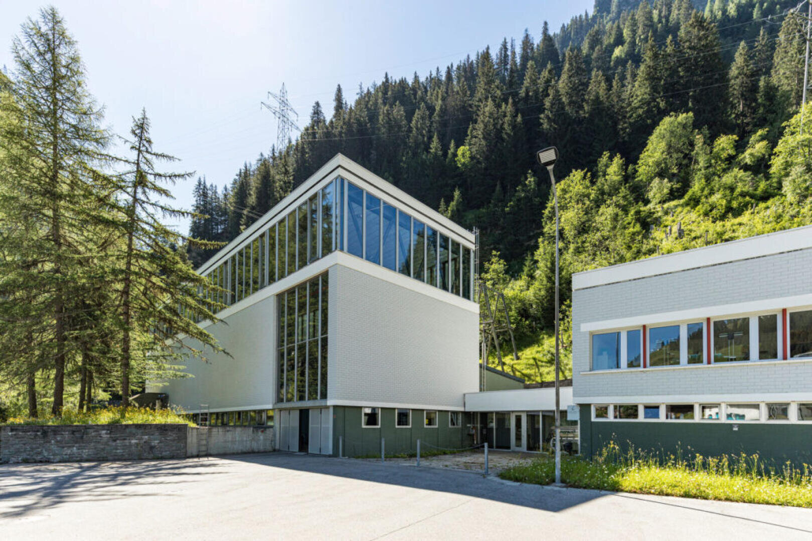 Ein modernes, weißes Gebäude mit großen Glasfenstern steht an einem sonnigen Tag am Fuße eines bewaldeten Hügels, umgeben von Bäumen und Grünflächen.