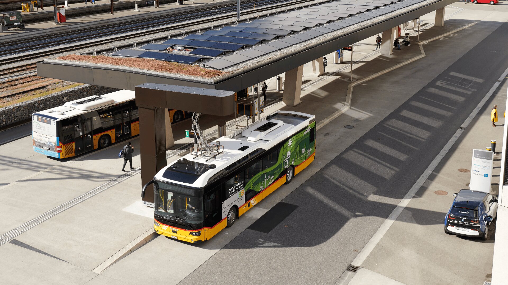 Ein gelber und grüner Elektrobus parkt an einer Ladestation unter einem Solarmodul-Dach, während in der Nähe an einem sonnigen Tag ein anderer Bus, Spaziergänger und Bahngleise zu sehen sind.