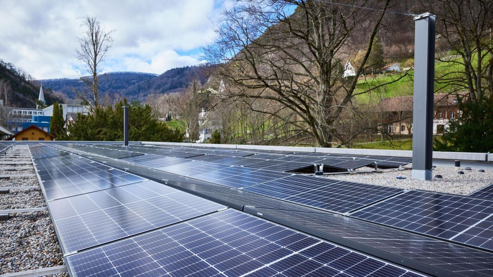 Reihen von Solarmodulen auf einem flachen Kiesdach mit Bäumen und grünen Hügeln im Hintergrund unter einem teilweise bewölkten Himmel.