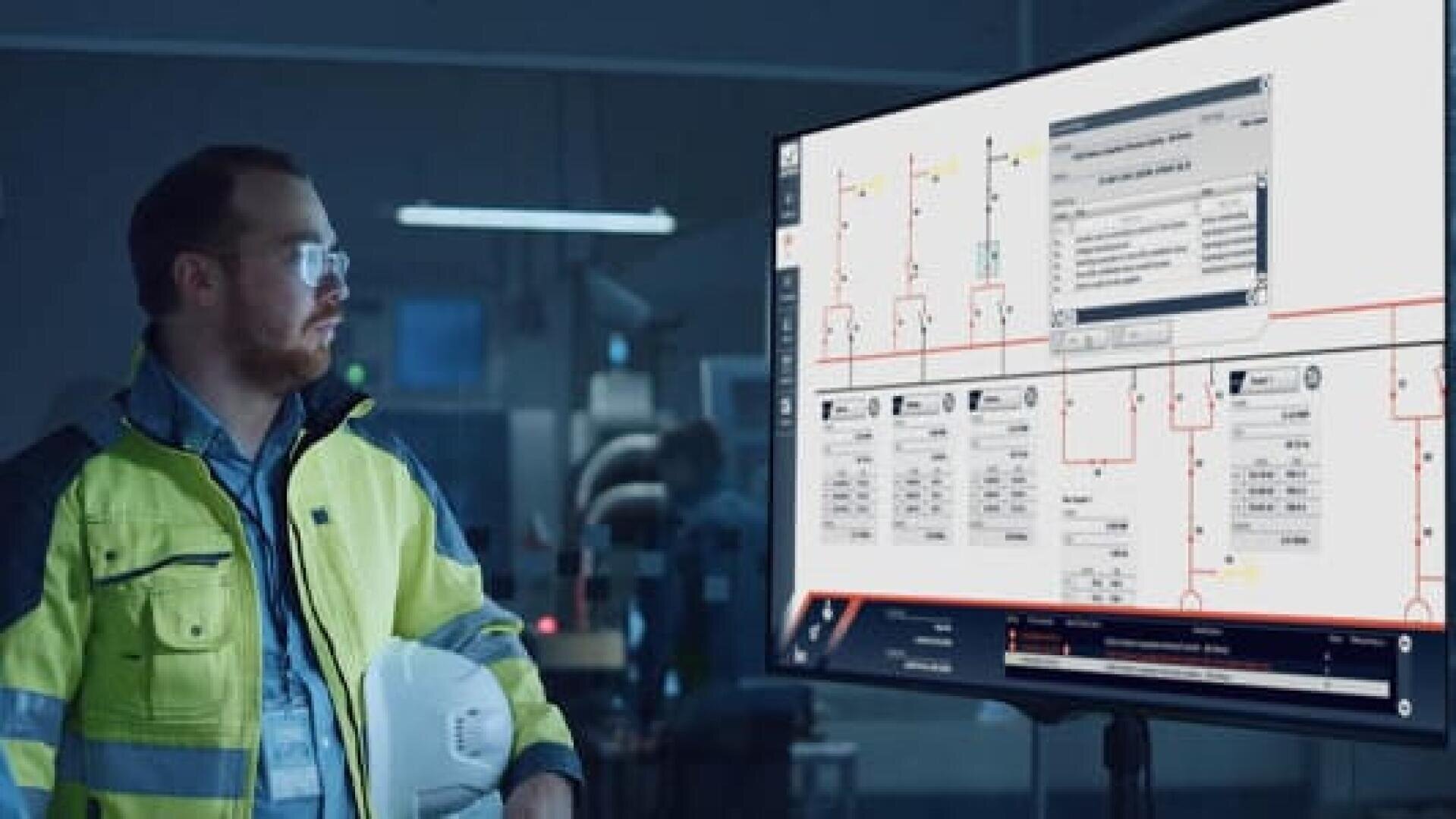 Ein Mann in Schutzkleidung und mit Brille steht mit einem weißen Schutzhelm in einem Raum und schaut auf einen großen Monitor, auf dem technische Diagramme und Daten angezeigt werden.