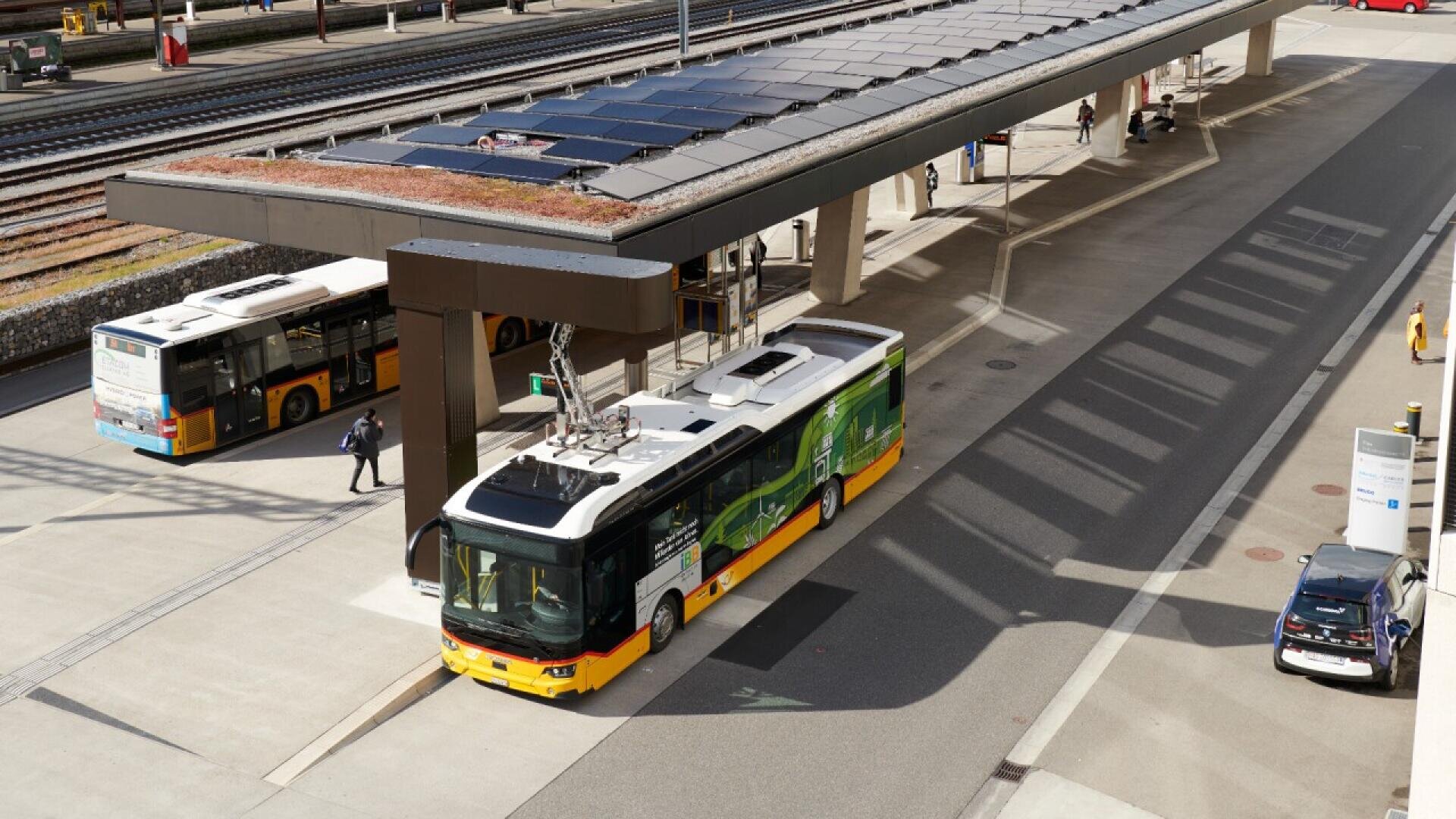 A yellow and green electric bus charges at a station under a solar panel roof. Another bus, people, and train tracks are visible nearby on a sunny day.