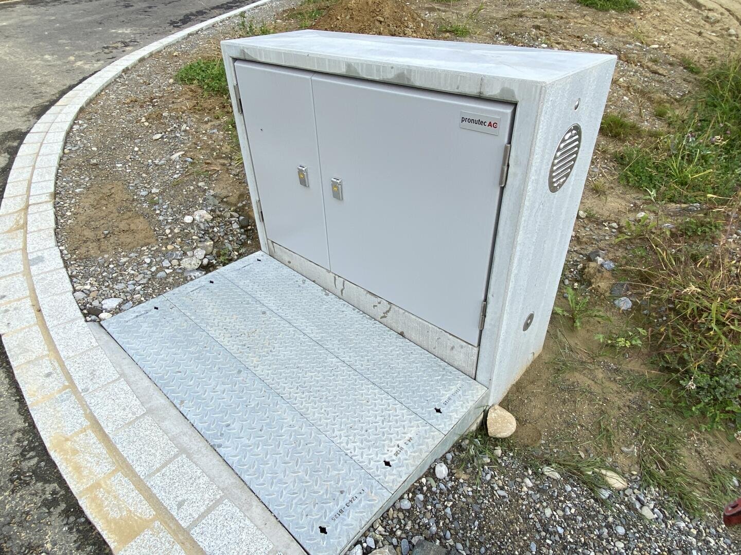 Ein großer grauer Metallkasten mit Doppeltüren und einer Entlüftungsöffnung steht neben dem Bürgersteig auf einem Fleckchen Erde und Schotter, davor eine Rampe aus Metallrauten.
