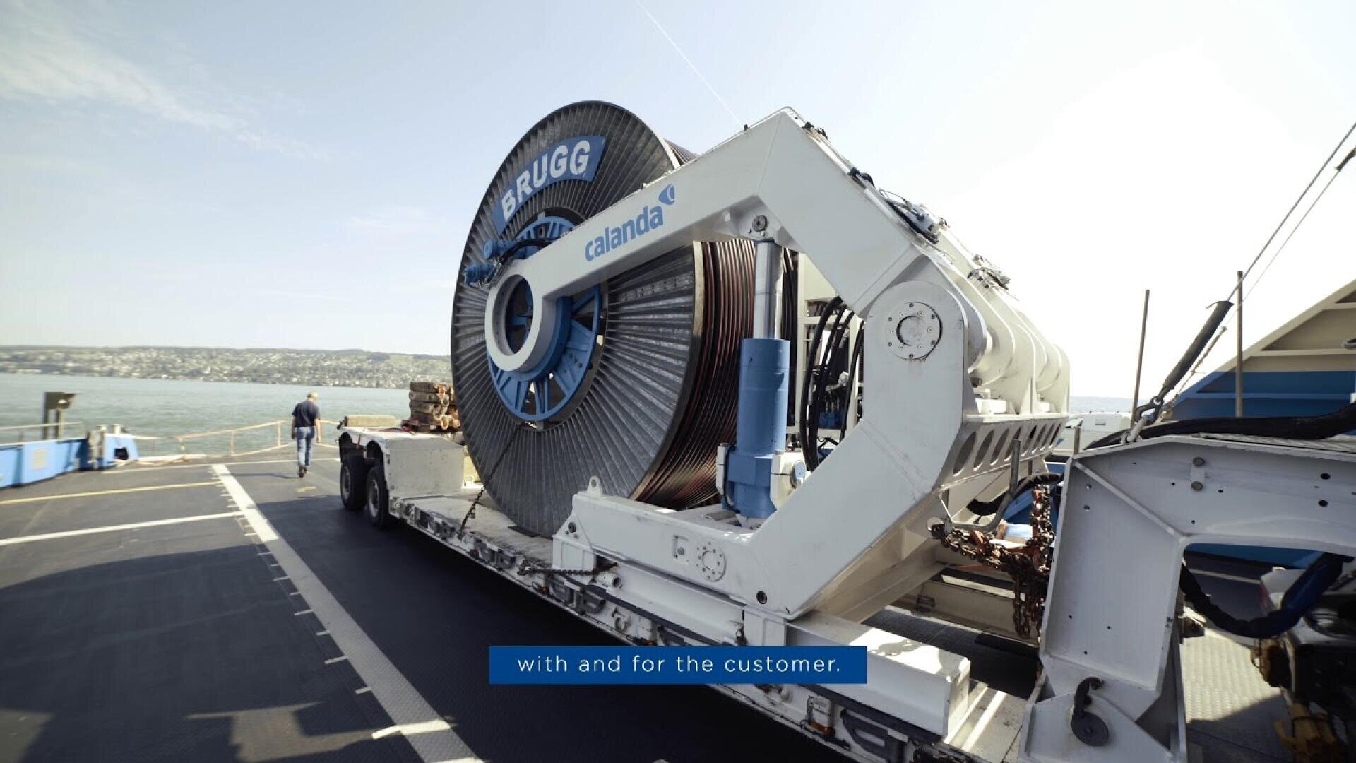 A large industrial cable spool labeled BRUGG and callanda is mounted on machinery on a docked vessel, with a body of water and distant shoreline in the background.