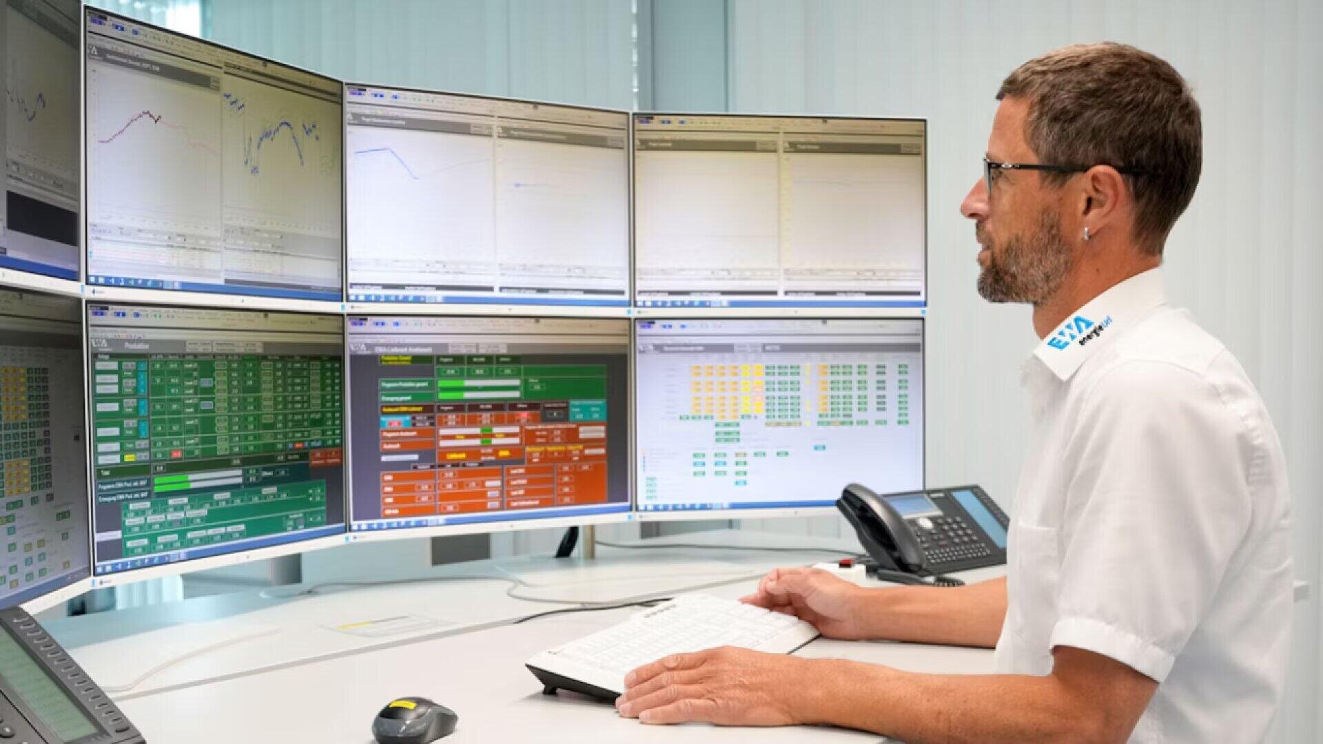 A man in a white uniform sits at a desk in front of six large computer monitors displaying charts, data tables, and graphs in a modern control room. He types on a keyboard and focuses on the screens.