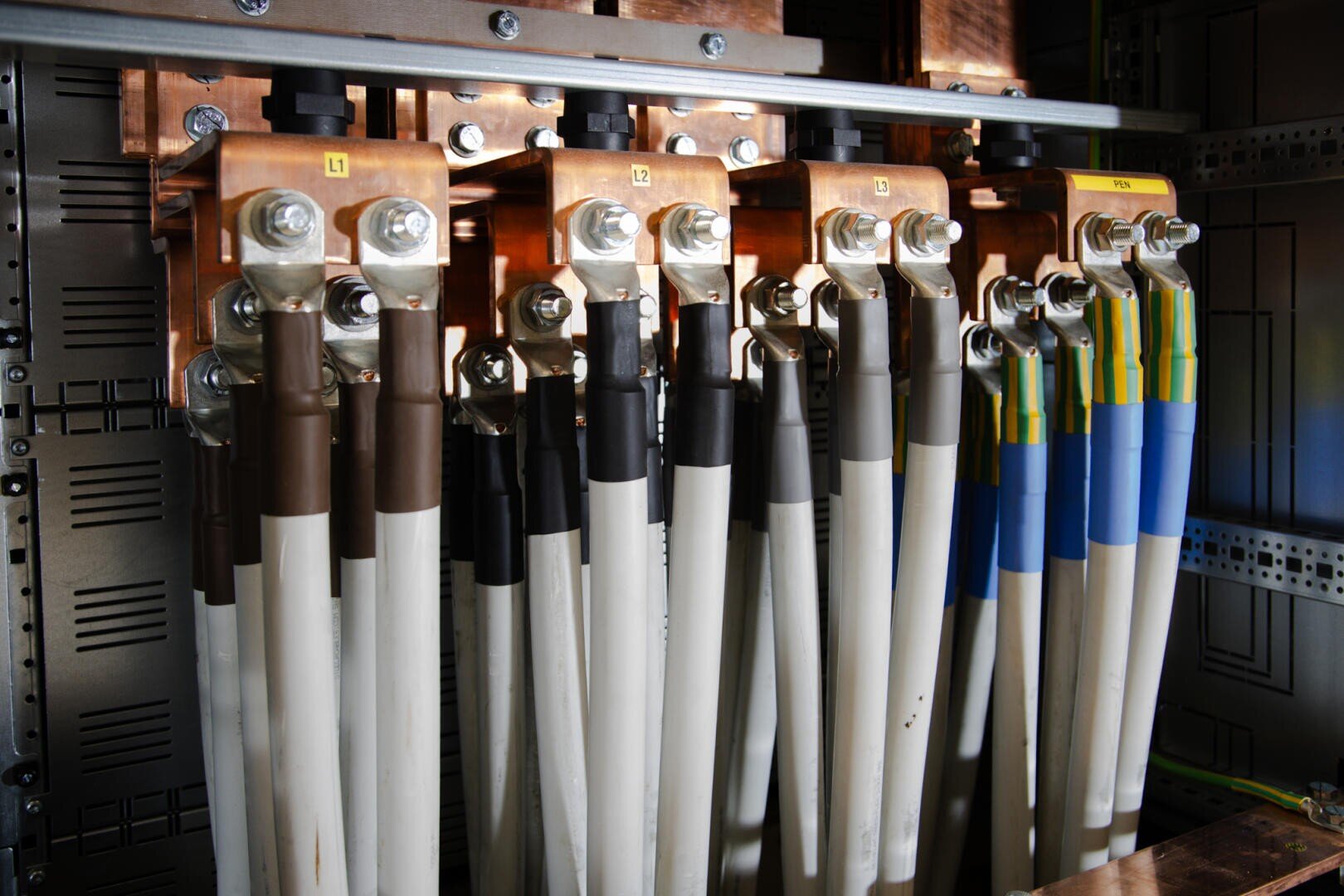 Nahaufnahme mehrerer dicker elektrischer Kabel, die an Kupferschienen in einer Schalttafel angeschlossen sind, wobei jedes Kabel farblich gekennzeichnet und beschriftet ist, was ein Stromverteilungssystem darstellt.