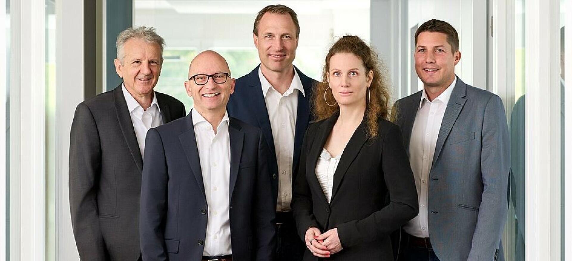 Fünf professionell gekleidete Personen, drei Männer und eine Frau, stehen in einem modernen Büro mit Glastüren und Fenstern zusammen, lächeln und schauen in die Kamera.