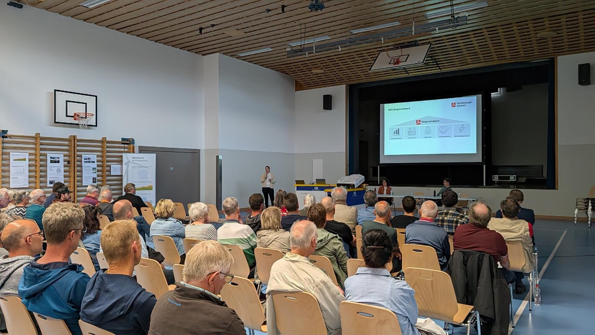 Eine Gruppe von Personen sitzt in einer Sporthalle vor einer Bühne, auf der eine Präsentation auf eine Leinwand projiziert wird. Ein Redner steht in der Nähe der Leinwand, und einige Teilnehmer machen sich Notizen.