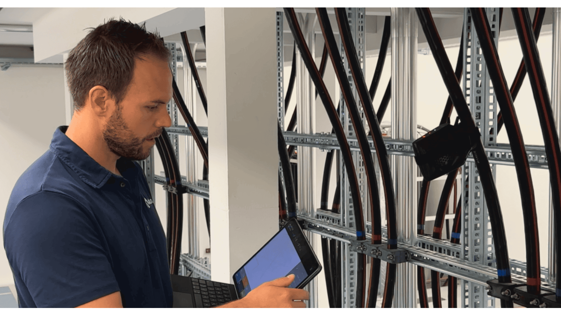A man in a blue shirt uses a tablet while inspecting large electrical cables organized on metal racks in a technical room.