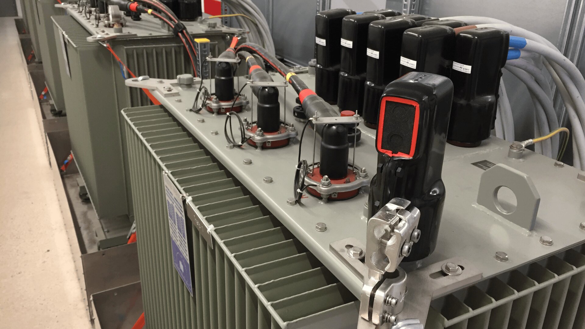 Nahaufnahme elektrischer Transformatoren in einem Kontrollraum, mit Verkabelung, Steckern und anderen Komponenten auf den Metallgehäusen.