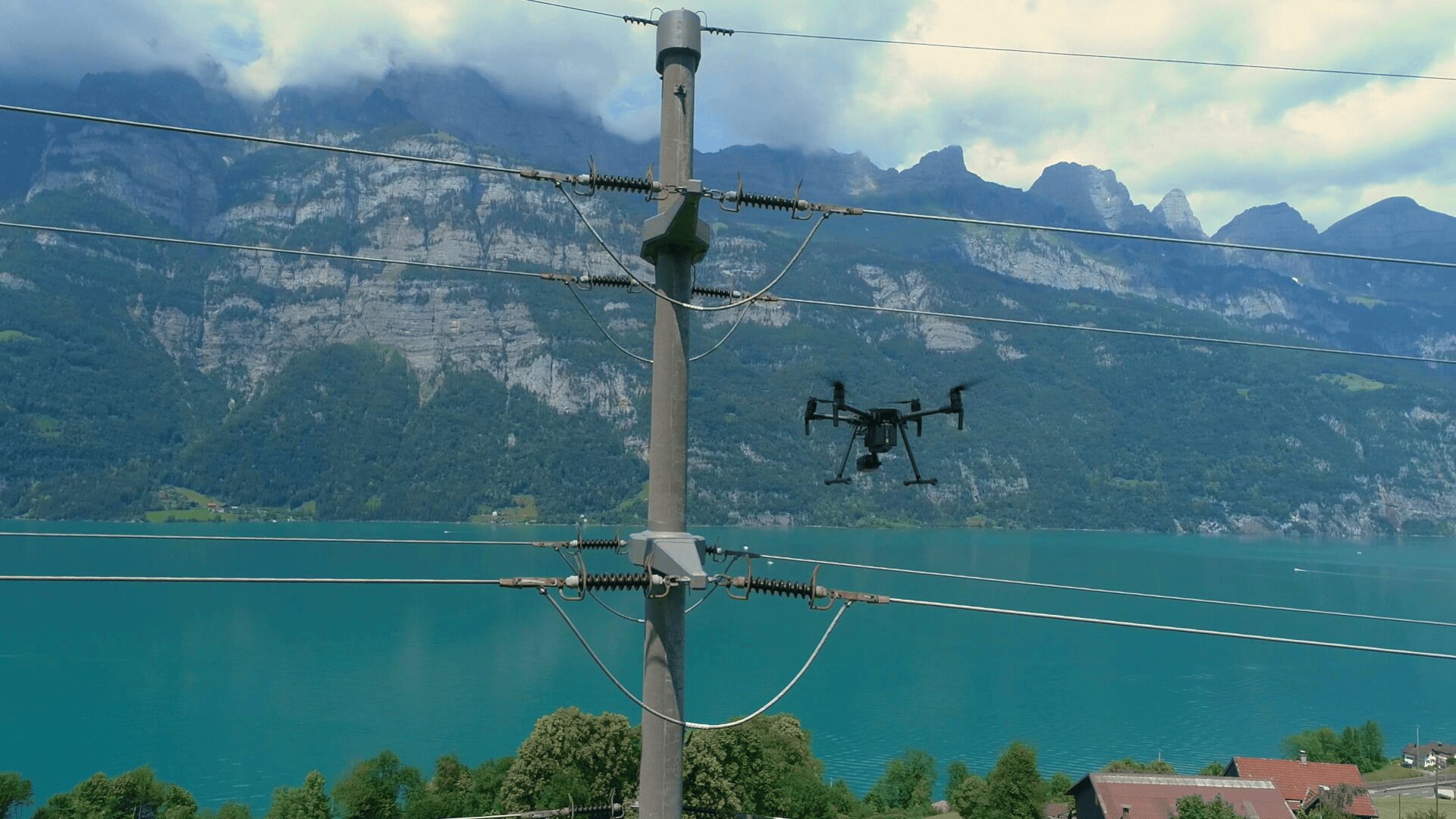 Eine Drohne fliegt in der Nähe von Stromleitungen über einem türkisfarbenen See, mit hohen Bergen und grünen Wäldern im Hintergrund. In der unteren rechten Ecke ist ein kleines Gebäude mit einem roten Dach zu sehen.