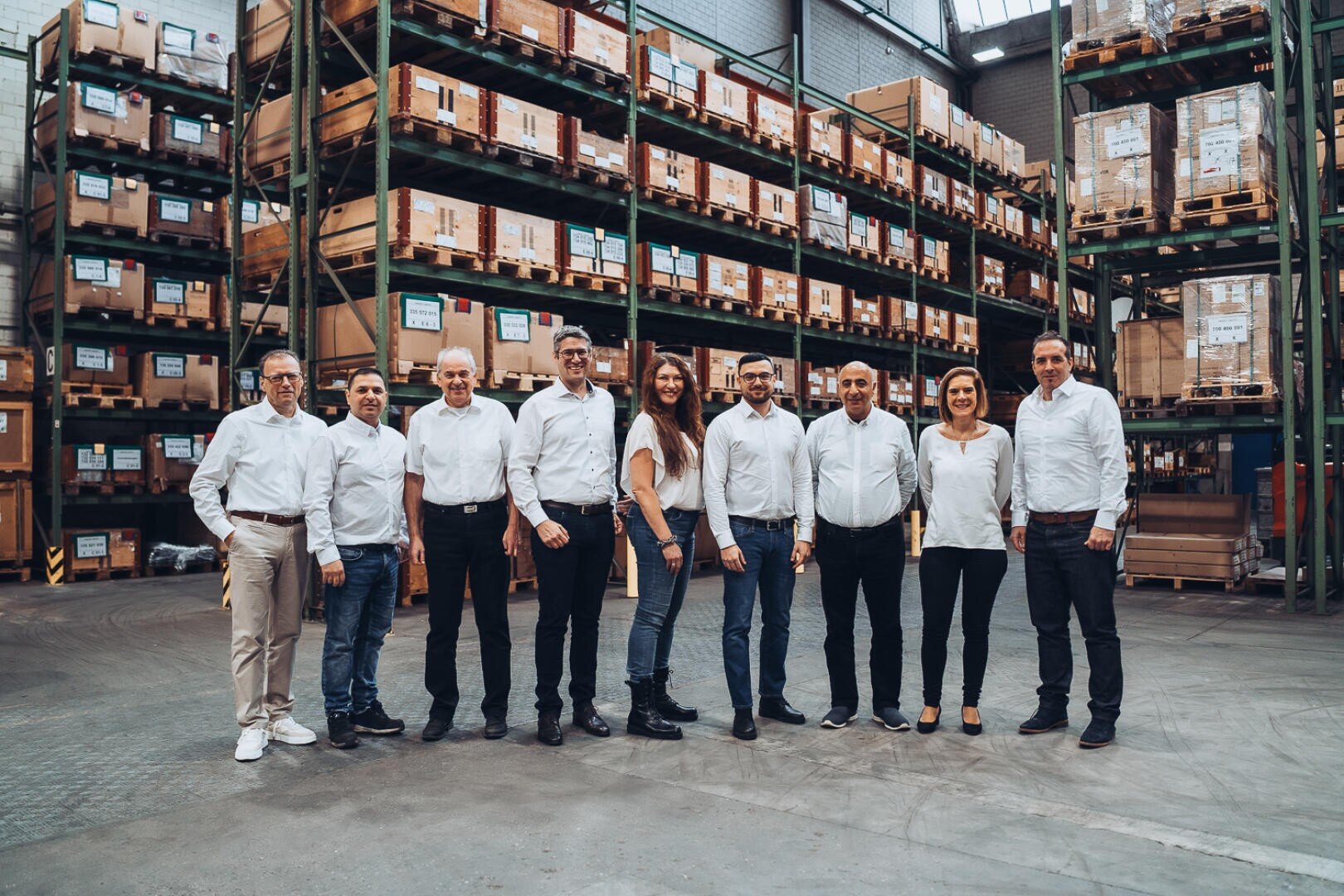Eine Gruppe von zehn Personen in legerer Business-Kleidung steht nebeneinander und lächelt in die Kamera. Sie befinden sich in einem Lagerhaus mit hohen Regalen, in denen sich Kisten und Paletten stapeln.