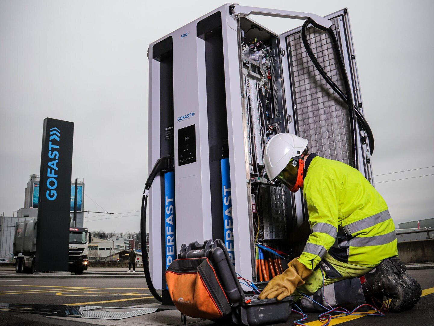 Ein Arbeiter mit gelber Schutzjacke und Helm repariert eine offene Ladestation für Elektrofahrzeuge mit Werkzeugen und Kabeln auf dem Boden, in der Nähe eines GOFAST-Schildes in einem Industriegebiet.