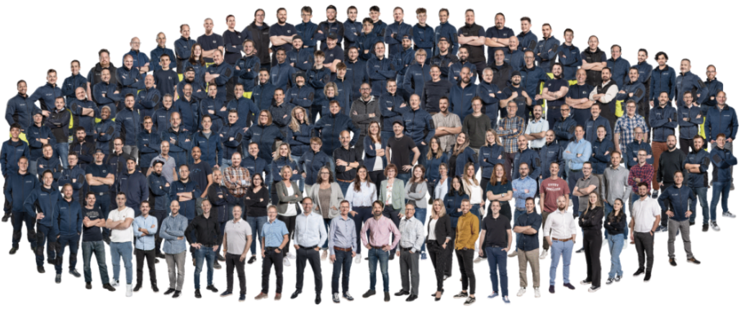 Eine große Gruppe von Menschen, meist Männer, stellt sich für ein Gruppenfoto vor einem weißen Hintergrund auf. Die meisten tragen marineblaue Hemden, einige tragen Freizeit- oder Geschäftskleidung in hellen Farben.