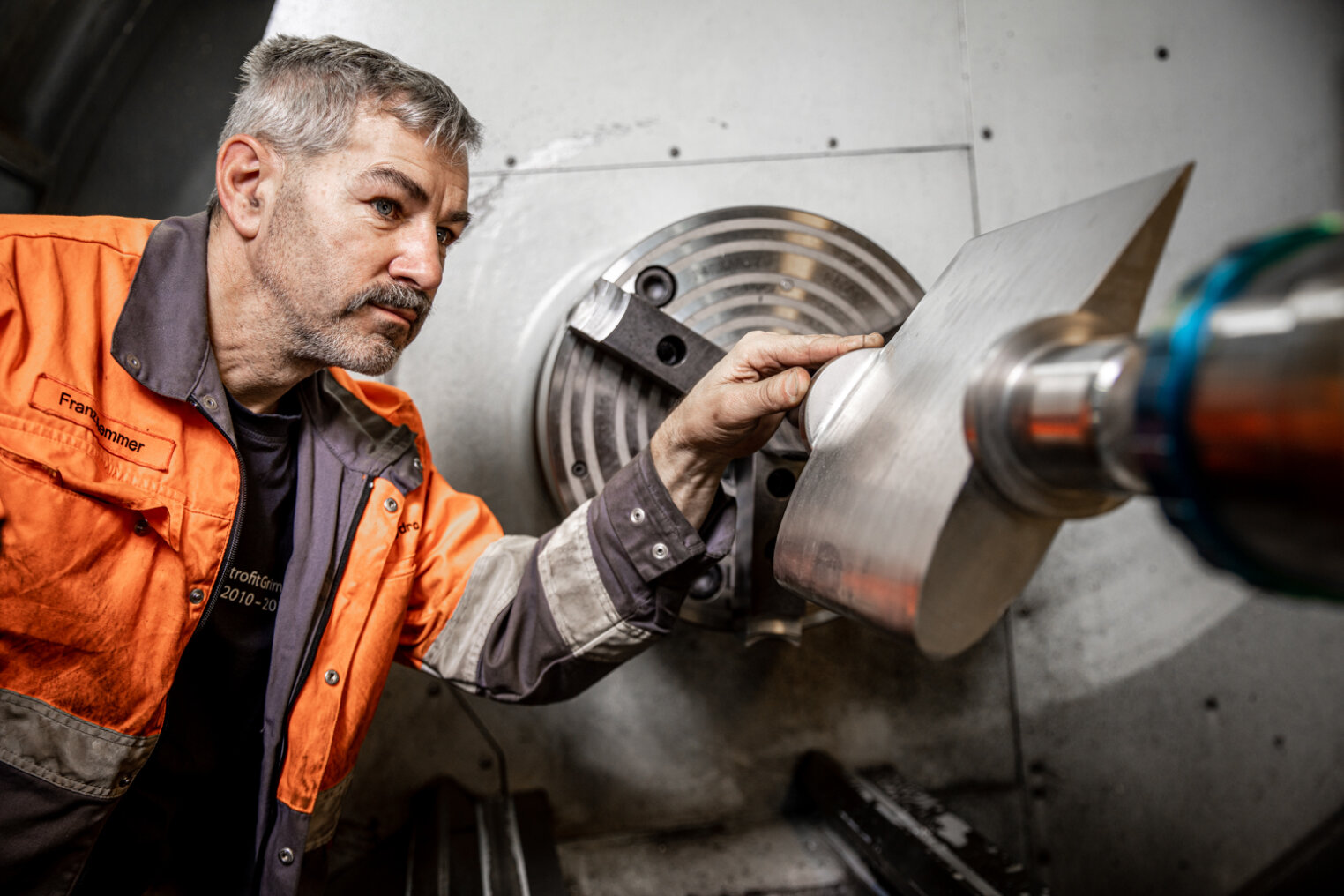 Ein Mann in einer orangefarbenen Arbeitsjacke prüft ein Metallteil, das in einer Drehmaschine in einer Industriewerkstatt gehalten wird, genau.
