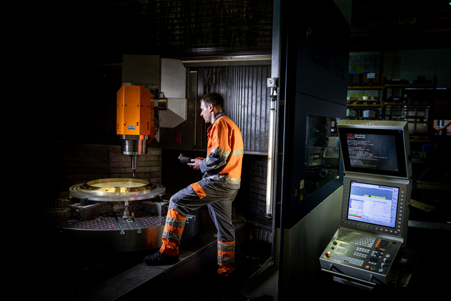 Ein Arbeiter in orangefarbener Schutzkleidung bedient in einer schwach beleuchteten Fabrik eine große CNC-Maschine, während im Vordergrund ein Bedienfeld Daten anzeigt.