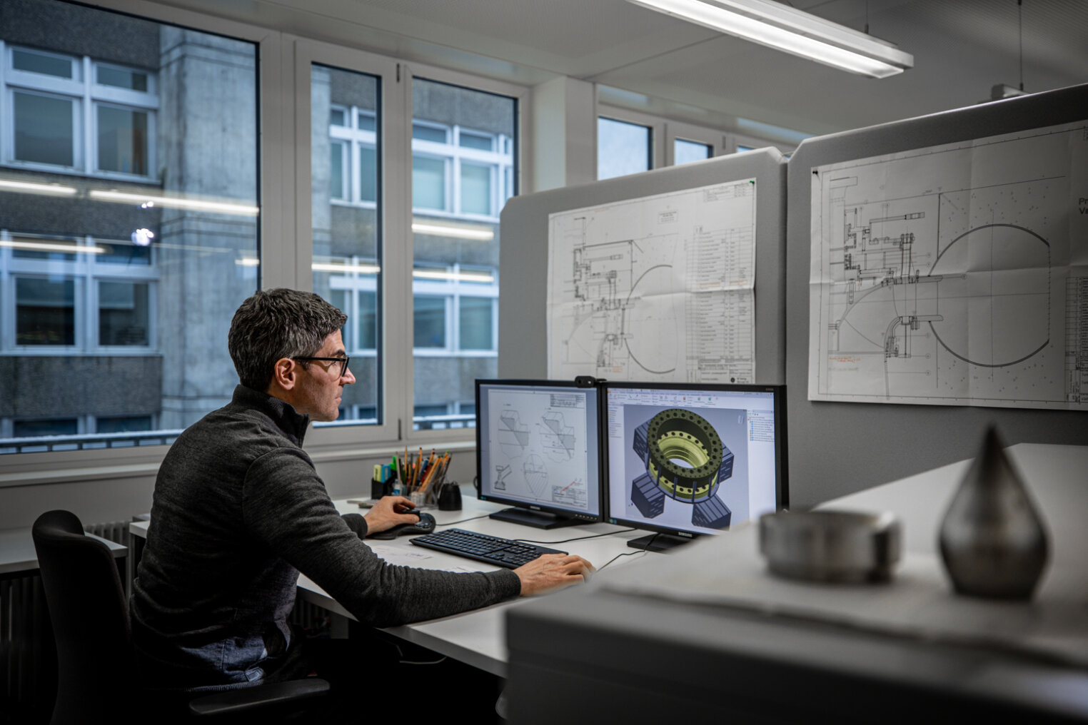 Ein Mann sitzt an einem Bürotisch und arbeitet an zwei Computermonitoren, von denen einer eine mechanische 3D-Konstruktion anzeigt. An den Wänden der Kabine sind technische Zeichnungen angeheftet, und große Fenster geben den Blick auf ein Gebäude frei.